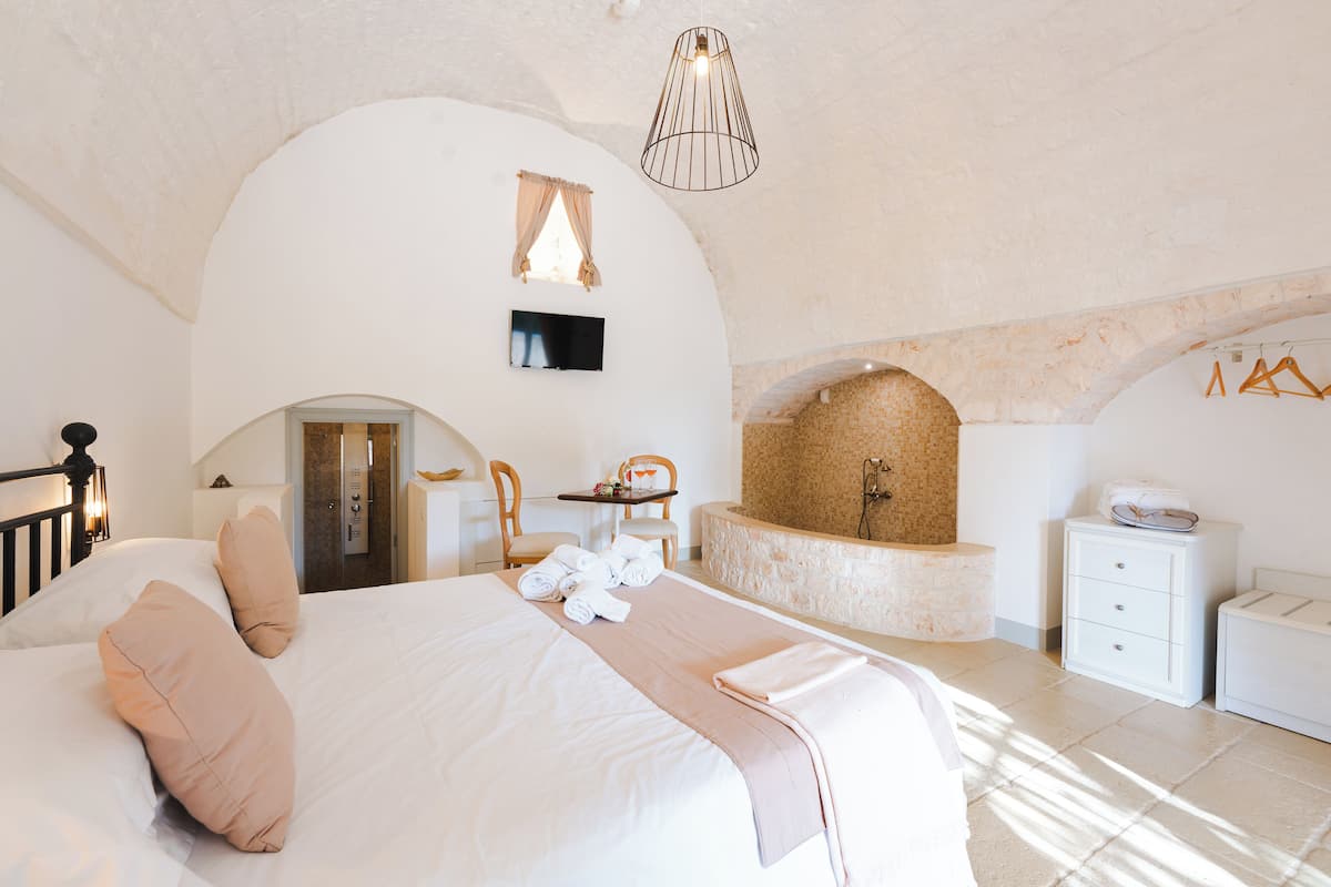 A bedroom with a large bed, a small table with chairs, a TV, a bathtub, and a wardrobe.