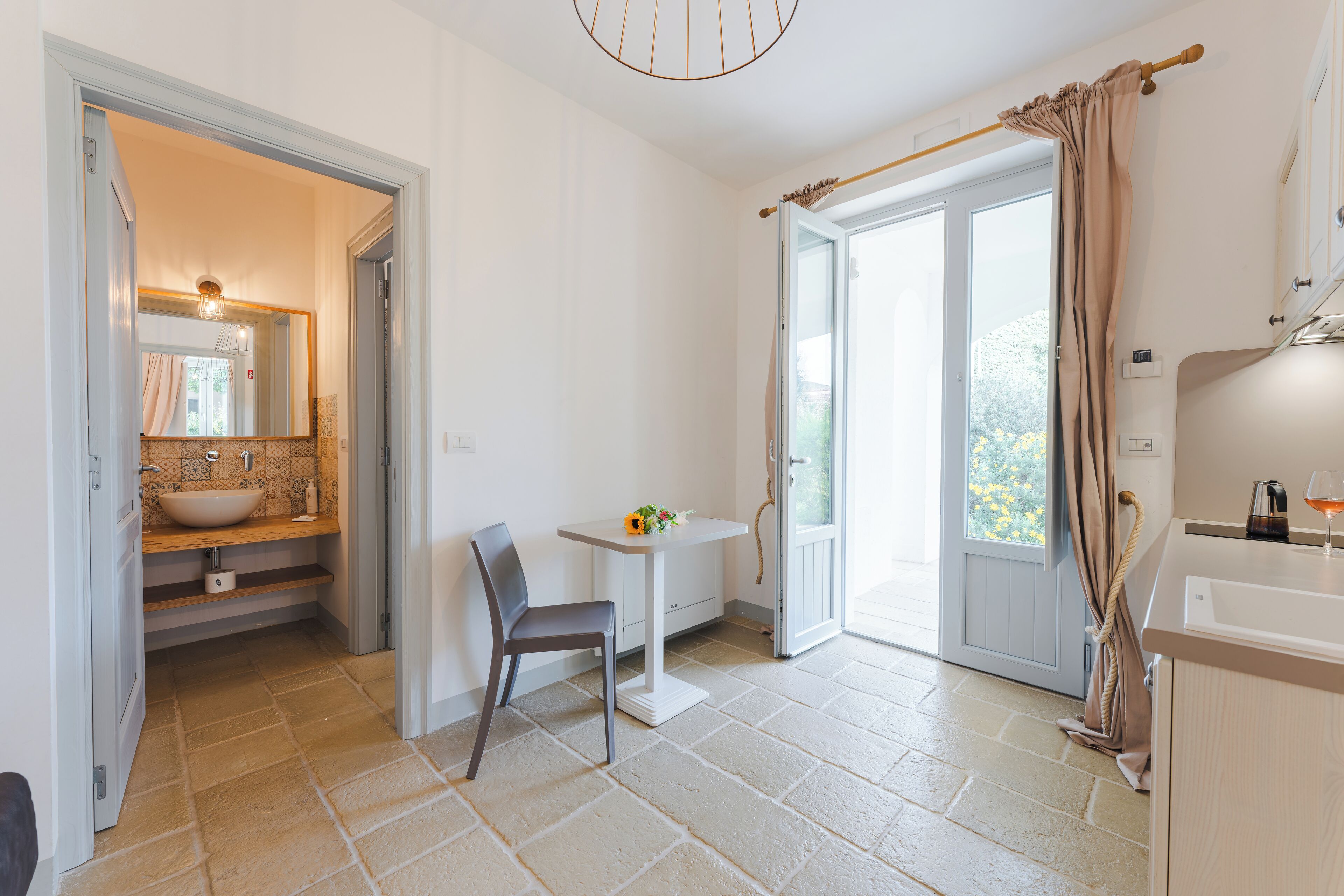 A modern bathroom with a sink, mirror, and shower. There is a chair and a small table with a vase of flowers.