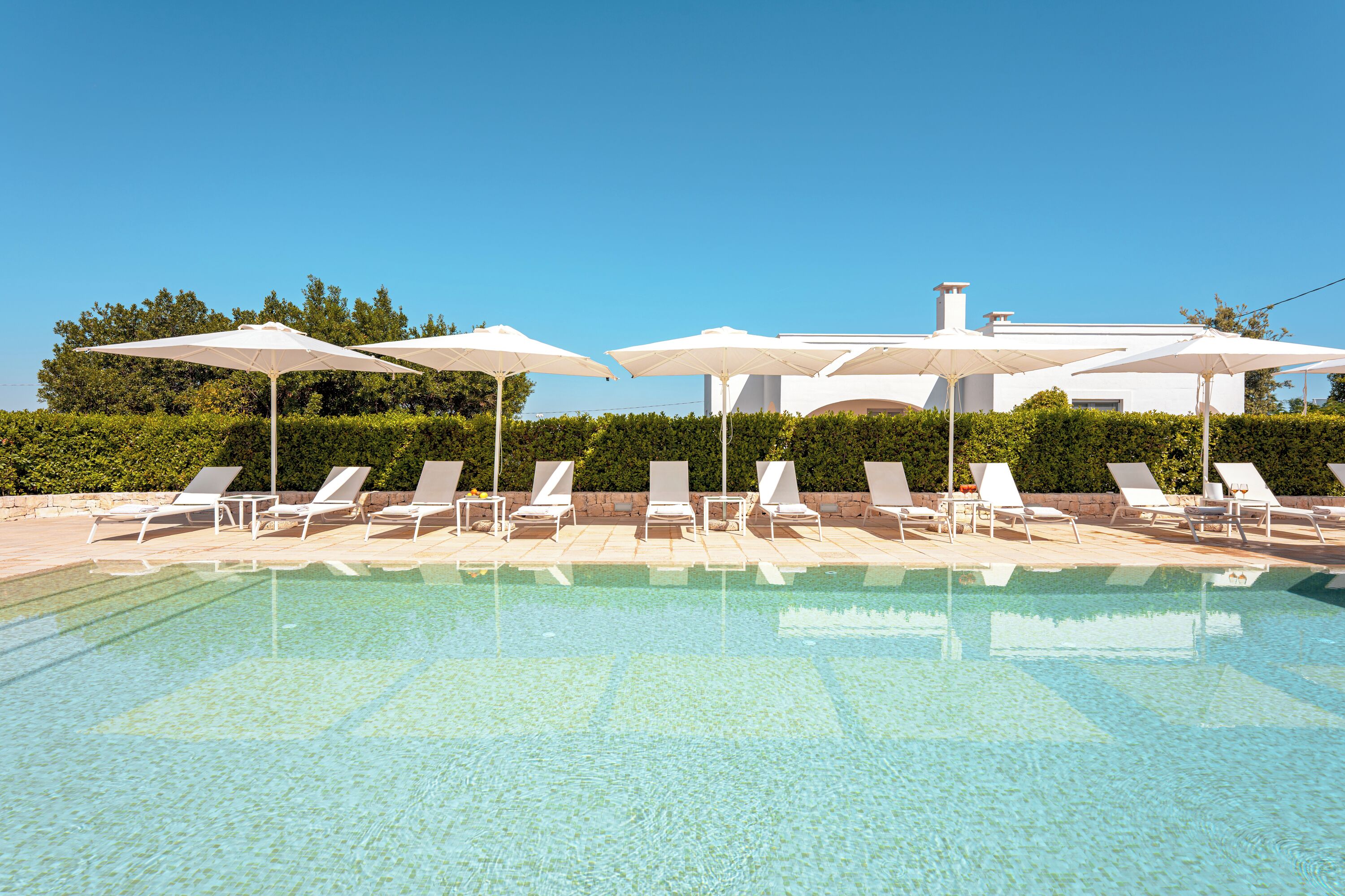 A clear swimming pool with white lounge chairs and umbrellas, surrounded by greenery and a building in the background.