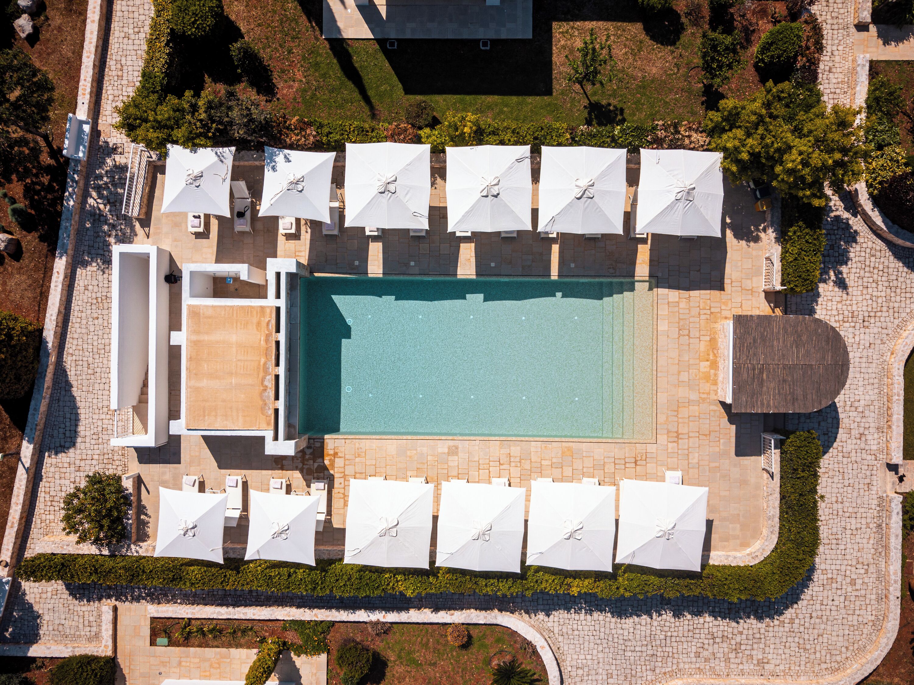 An aerial view of a hotel with a swimming pool, multiple white umbrellas, and a well-maintained garden.