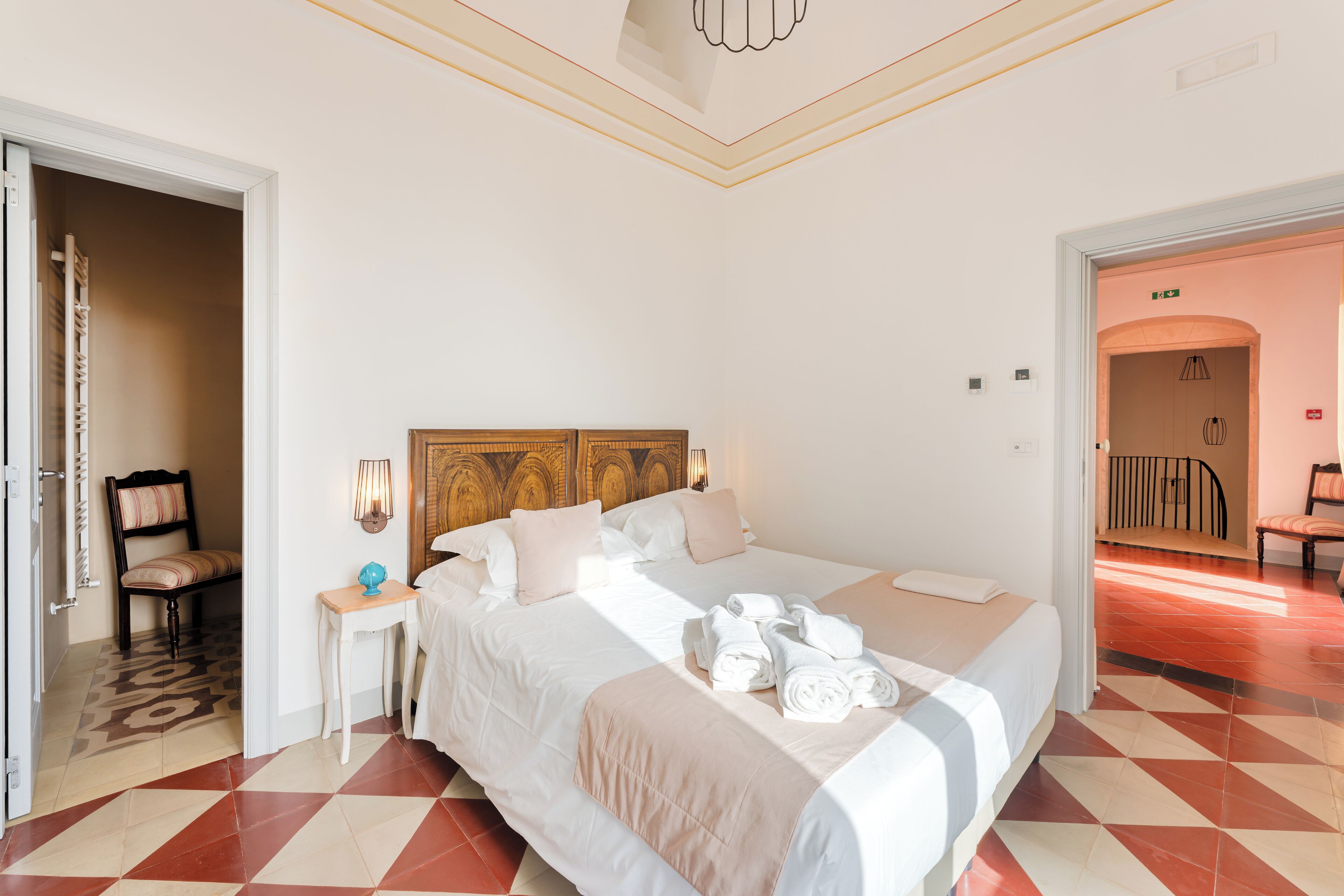 Quarto de hotel com uma cama grande, uma mesa pequena com uma lâmpada e uma cadeira. Há uma porta que dá para outro quarto.