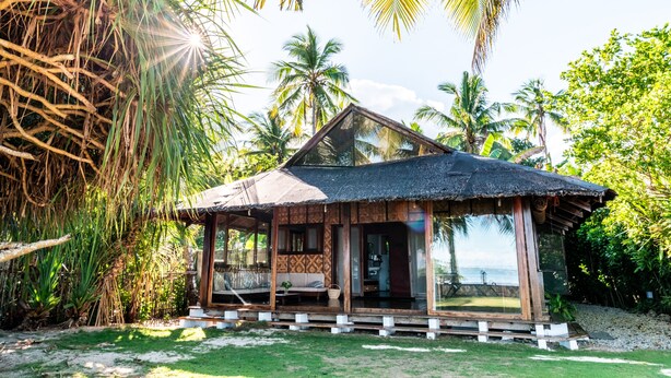 A small, thatched-roof hut with a porch, surrounded by lush greenery and palm trees, overlooking a body of water. - Greenhouse Siargao (General Luna)