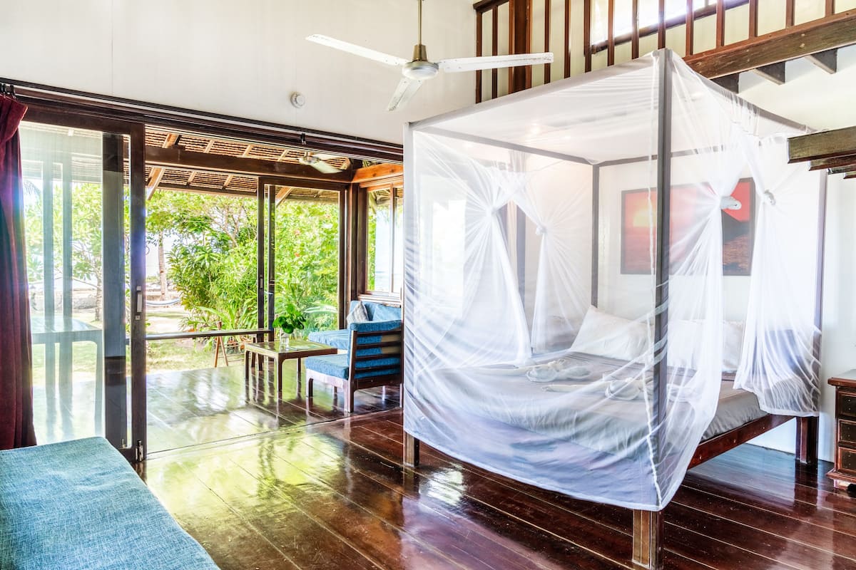 A room with a bed under a mosquito net, a ceiling fan, and a view of greenery outside.