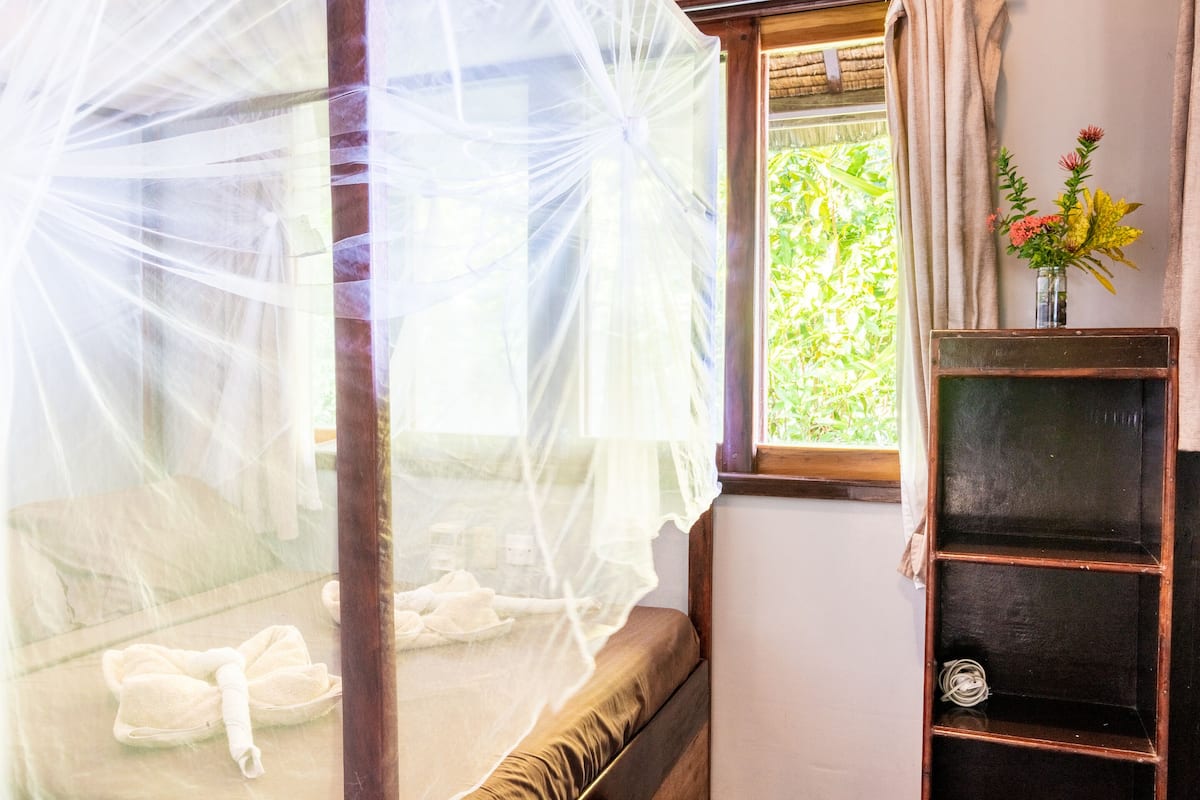 A bedroom with a mosquito net over the bed, a wooden headboard, a bedside table with a vase of flowers, and a window with a view of greenery.