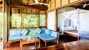 A wooden room with a blue sofa, a wooden coffee table, and a bed with a mosquito net. - Greenhouse Siargao (General Luna)