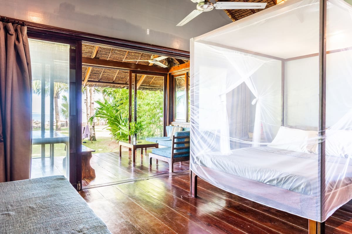 A room with a bed covered in a mosquito net, a wooden floor, a table with chairs, and a view of greenery outside.