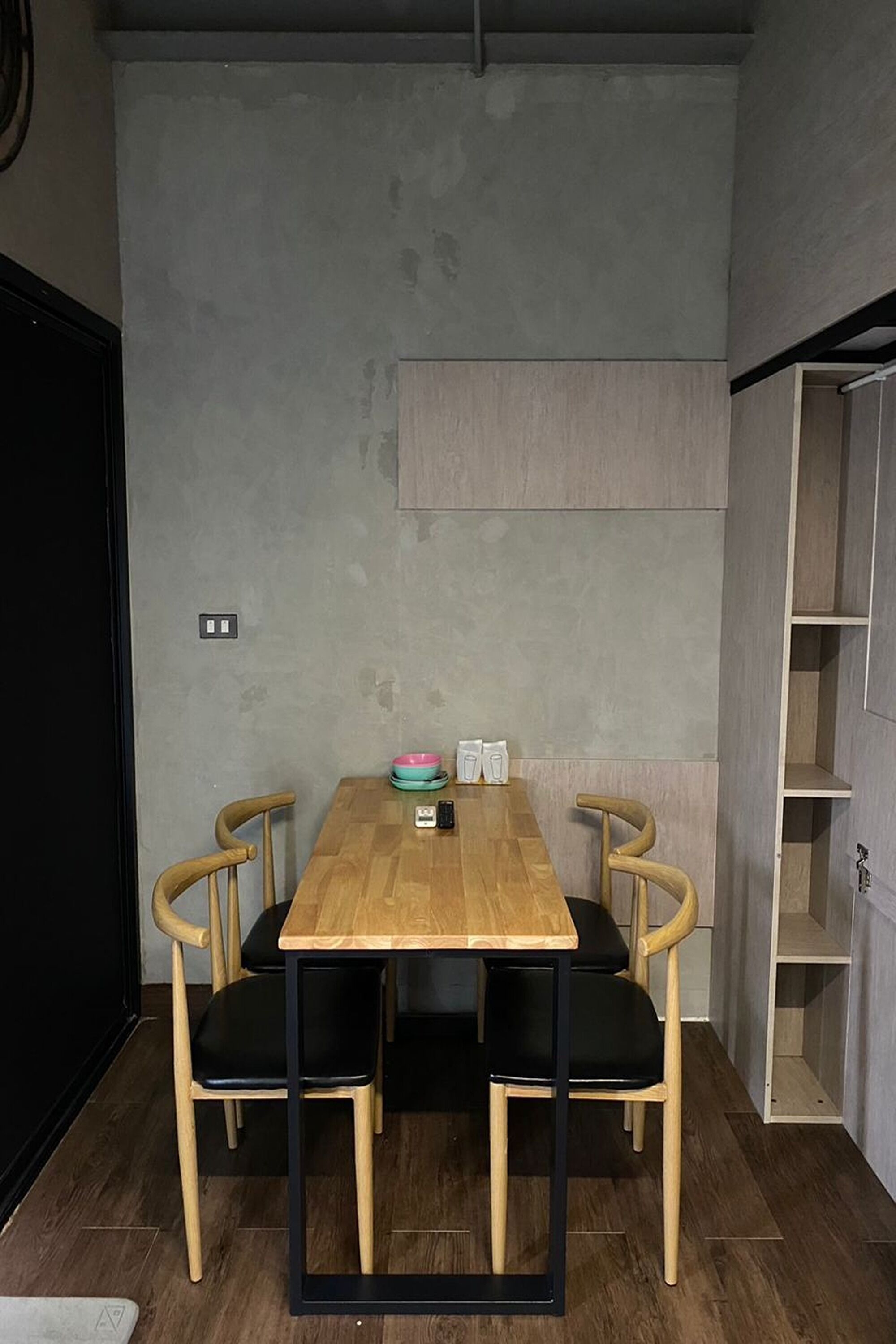 Un rincón comedor pequeño con una mesa de madera y sillas negras, un estante bajo y una pared lisa con un interruptor de luz.