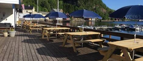 A wooden deck with picnic tables and blue umbrellas, overlooking a body of water with boats and a forested hill in the background.