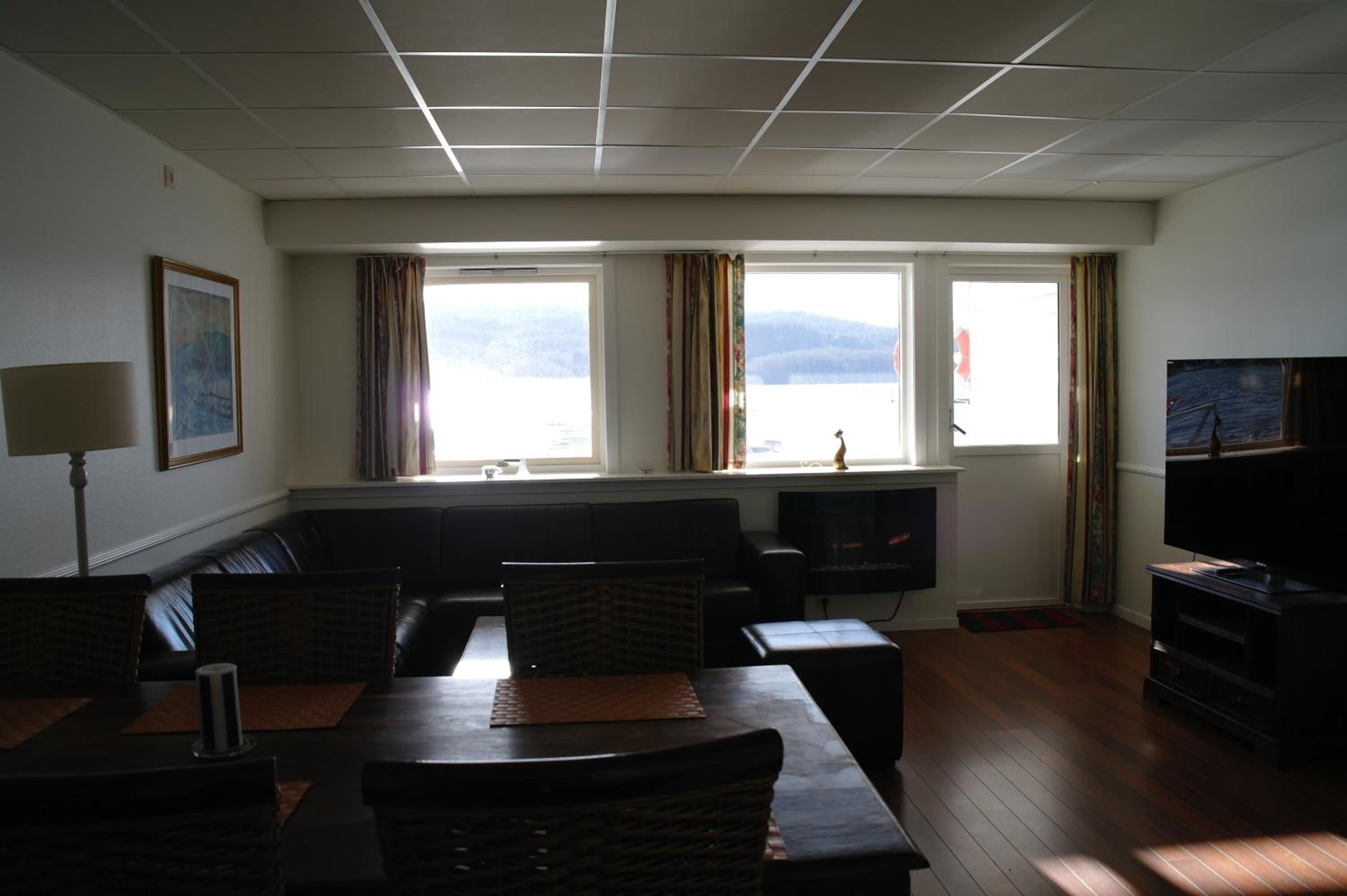 Una sala de estar con sofá, mesa de comedor y televisor. Hay dos ventanas con cortinas, chimenea y un cuadro en la pared.