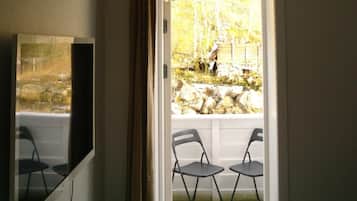 A room with a television mounted on the wall, a door leading to an outdoor area with two chairs and a stone wall.