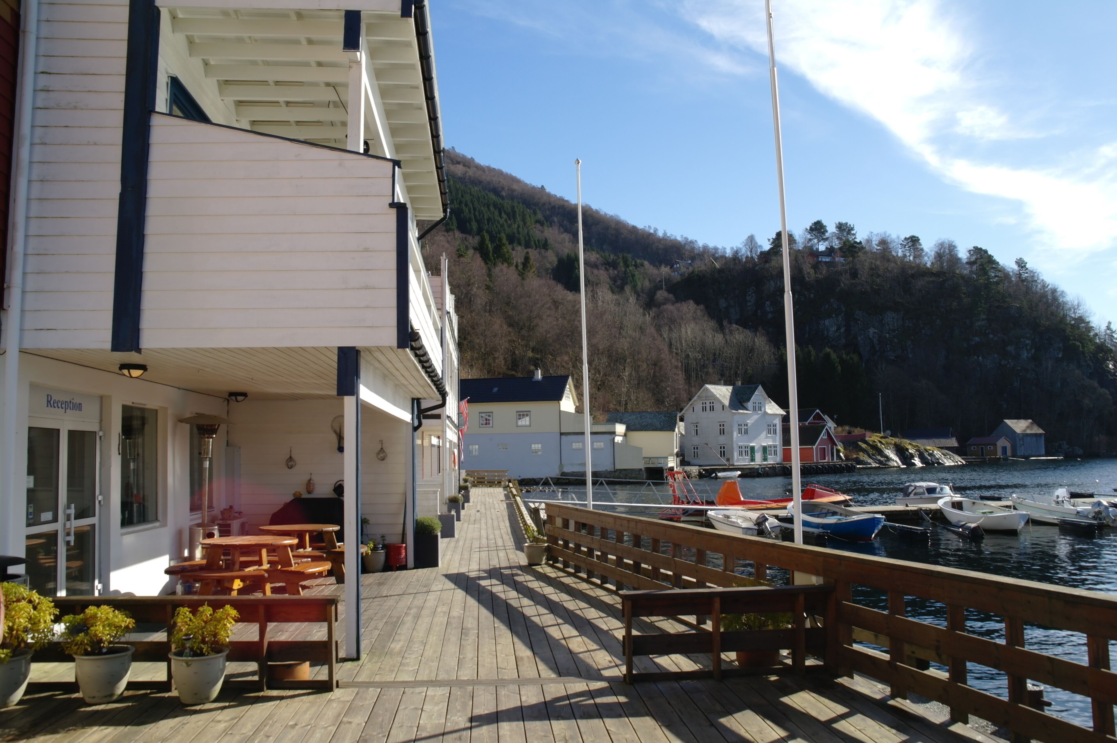 A wooden pier with outdoor seating, a reception area, and a view of boats and hills in the background.