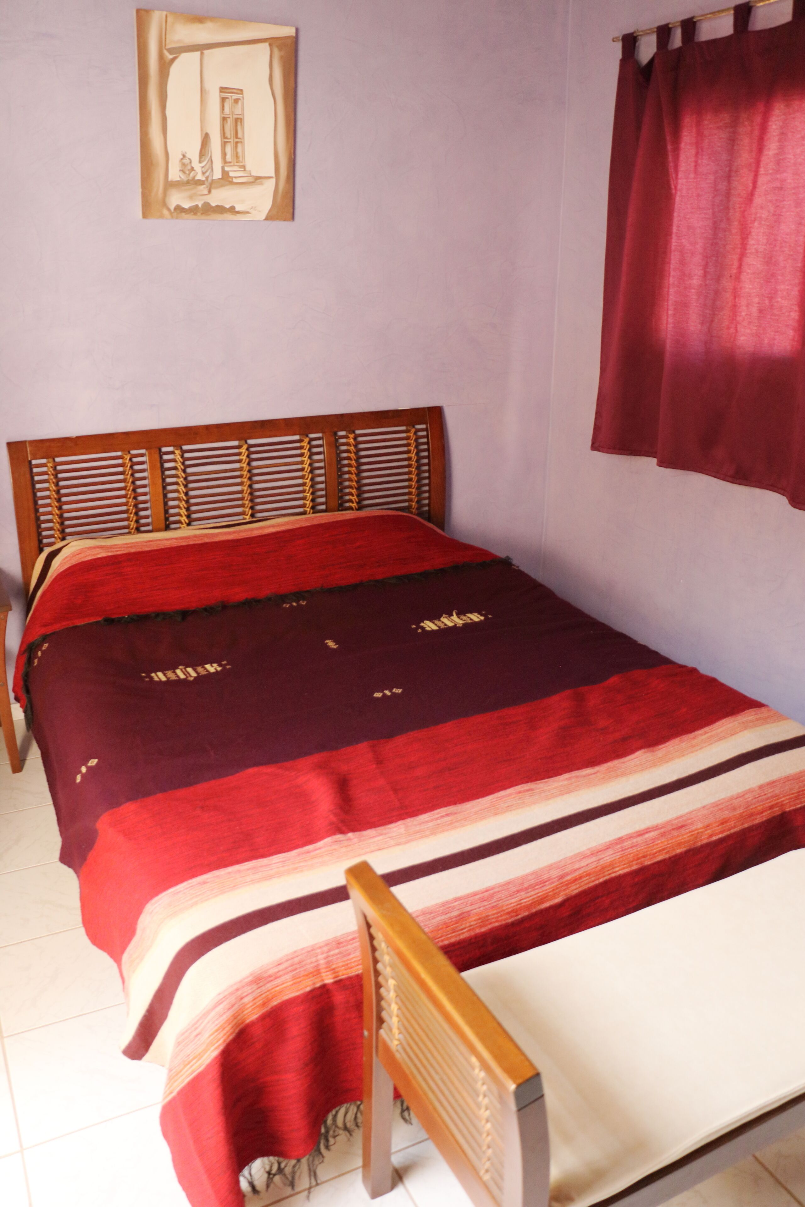 Una cama individual con cabecera de madera, una colcha de color rojo y beige, y una cortina roja en la ventana.