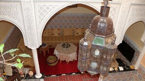 Un intérieur marocain traditionnel, avec des arches richement décorées, une lanterne suspendue, un tapis à motifs et une petite table sur laquelle se trouve une théière.
