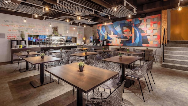 Interior moderno de restaurante con varias mesas y sillas para comer, una zona de cocina y un mural abstracto y colorido en la pared.