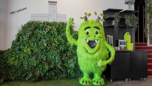 Un personaje mascota verde frente a un muro con ilustración de un paisaje urbano y un seto verde.