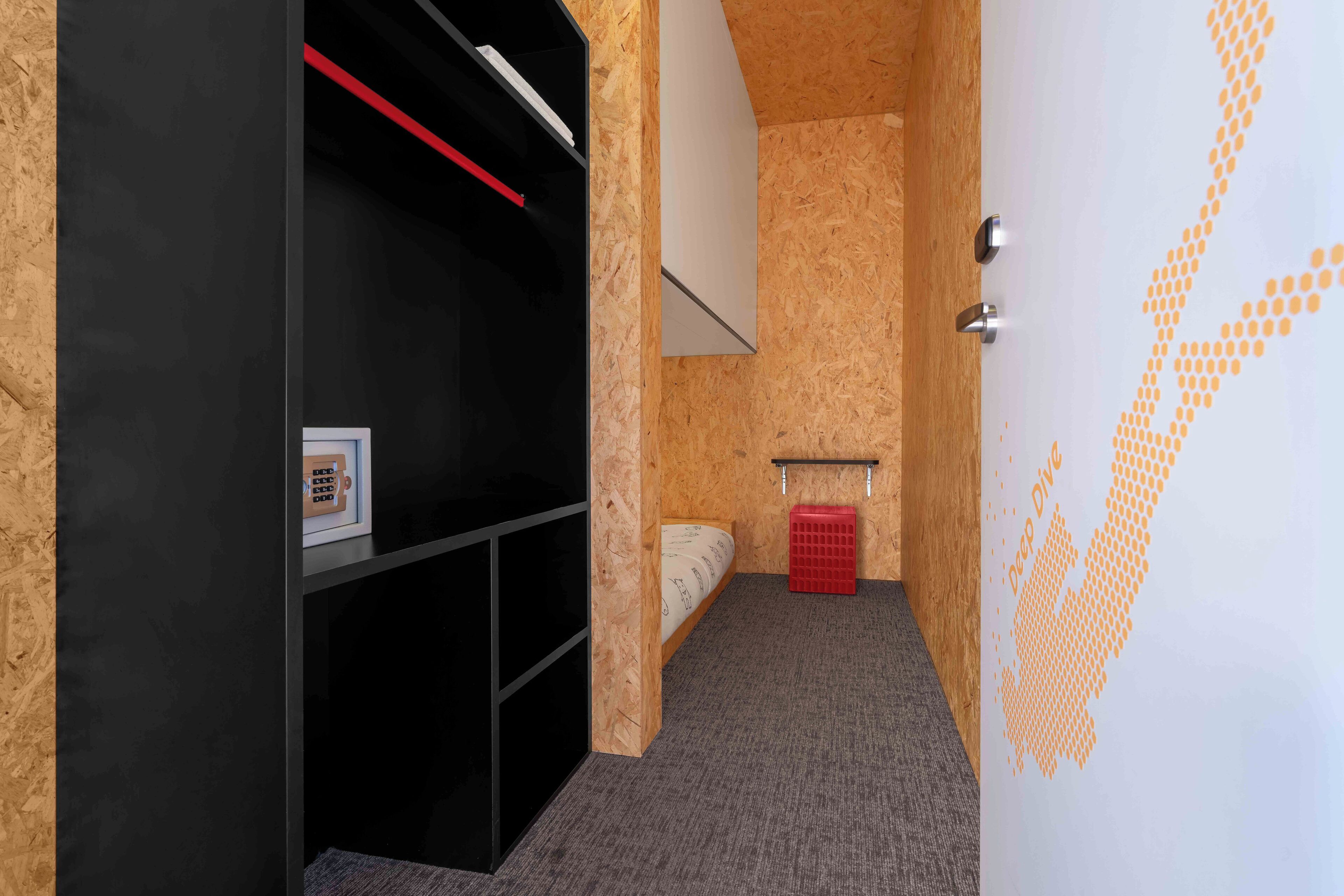 A compact room with a bed, a black wardrobe, a red basket, and a white door with a geometric pattern.