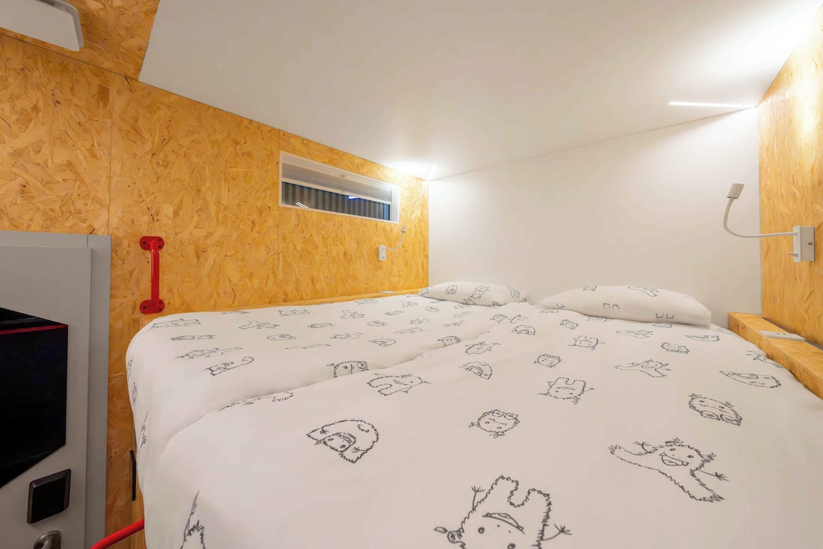 Ein Schlafzimmer mit einer Holzwand, einem Bett mit weißer Bettwäsche mit Tiermotiven, einem roten Gegenstand an der Wand und einem kleinen Fenster mit Lüftung.