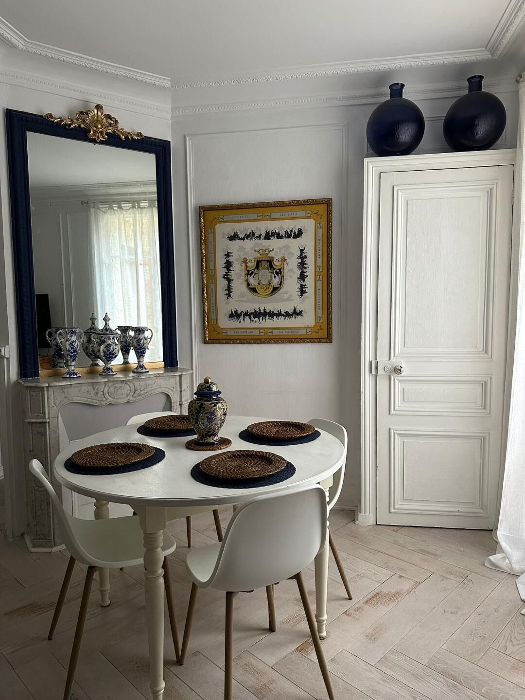 Zona pranzo con tavolo bianco, quattro sedie, uno specchio decorativo, un'opera d'arte incorniciata e una porta bianca.