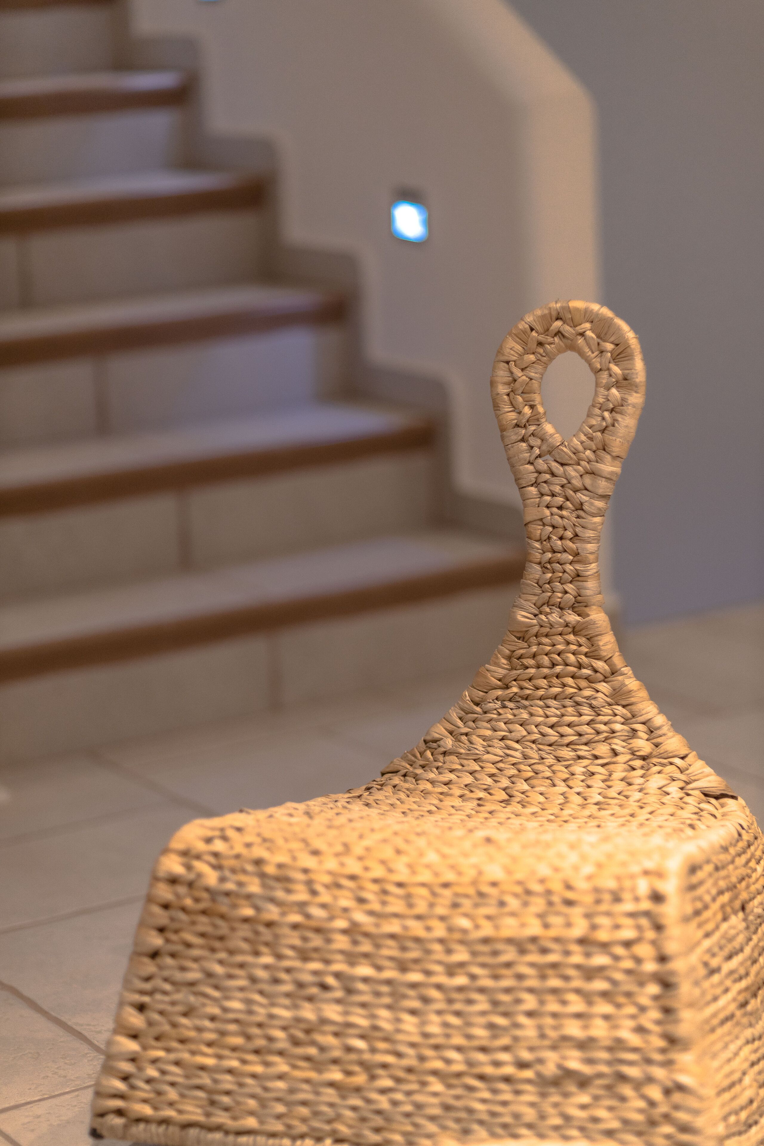A woven basket-like object with a handle, placed on a tiled floor near a staircase.
