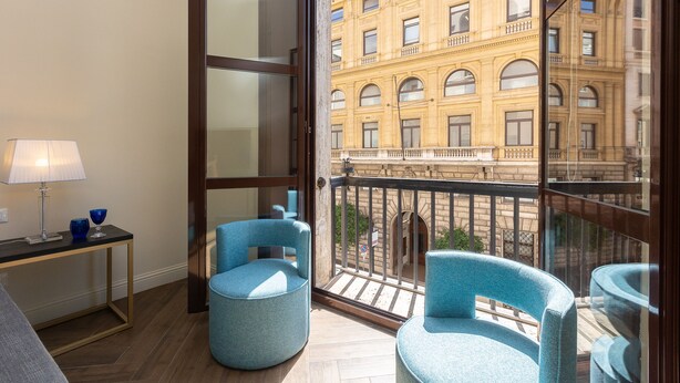 Een balkon met twee blauwe stoelen, een kleine tafel met een lamp en uitzicht op een geel gebouw.