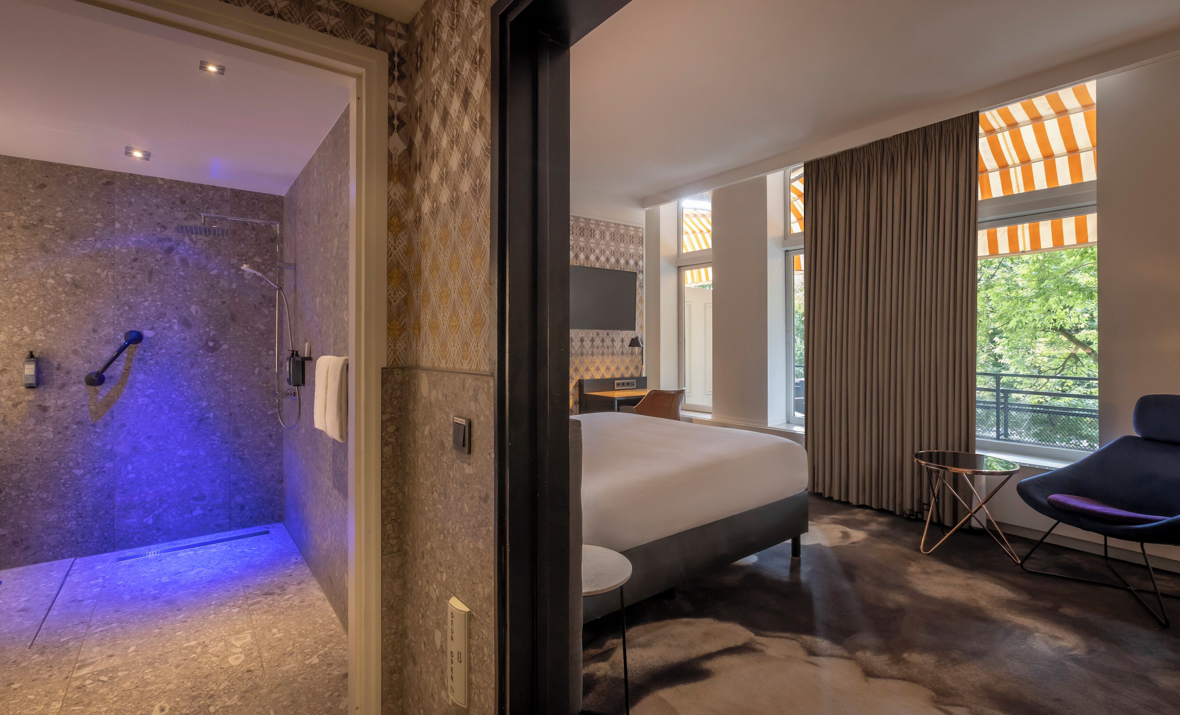 Ein modernes Hotelzimmer mit einer Duschkabine aus Stein, einem Bett, einem Schreibtisch und Blick auf Grünanlagen durch große Fenster.