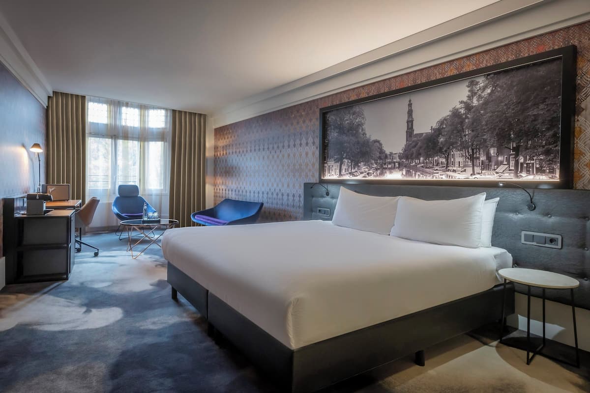Une chambre d’hôtel avec un grand lit, un bureau avec une chaise, une petite table et une grande fenêtre avec des rideaux.