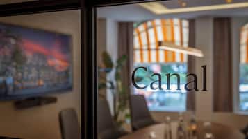 Vista a través de una ventana de un comedor con una mesa preparada para cuatro personas, un cuadro en la pared y la palabra "Canal" a la vista.