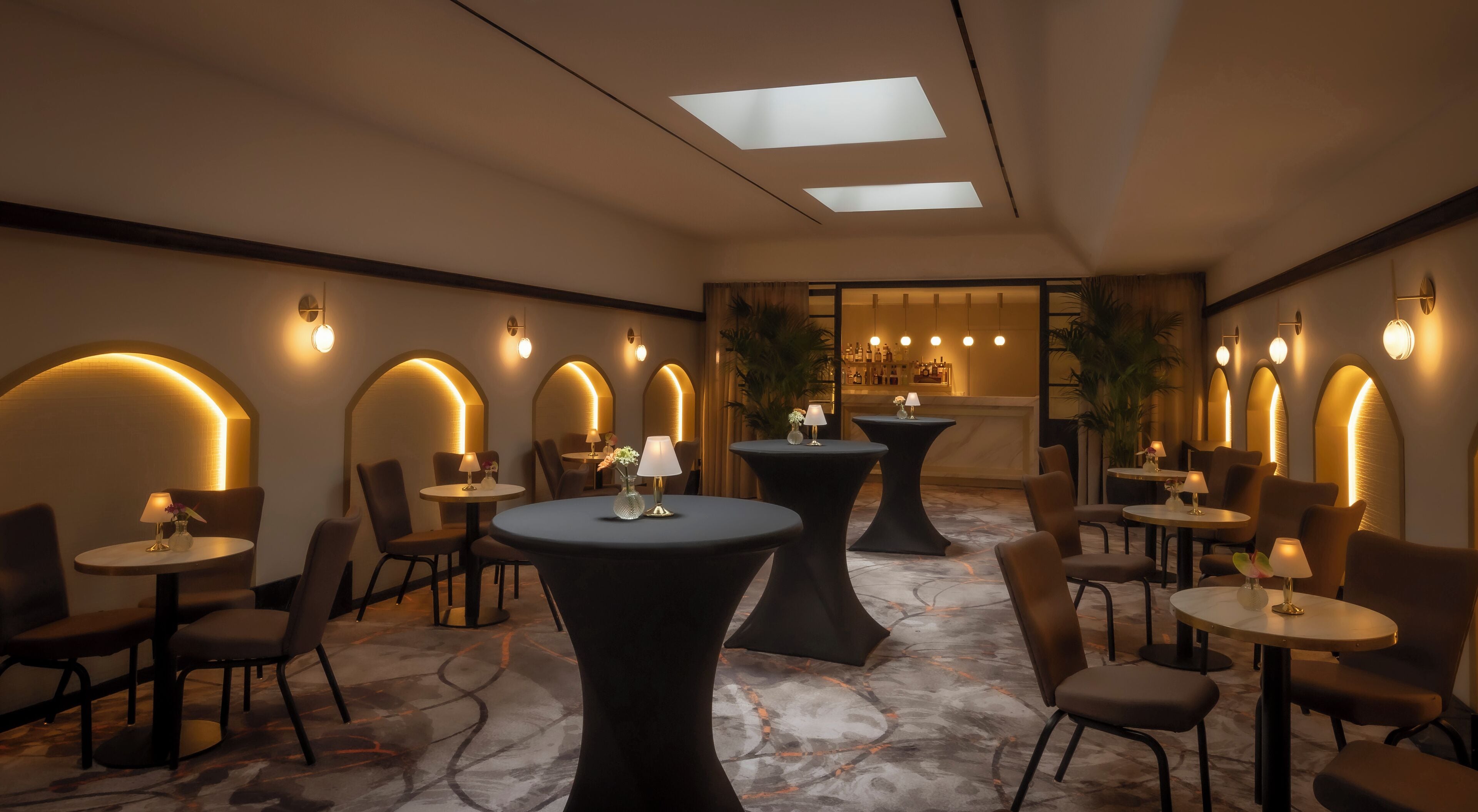 A modern lounge area with round tables and chairs, recessed lighting, and a view of a bar area through an arched doorway.