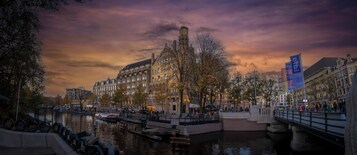 Un edificio histórico con una torre destacada, un canal con barcos y un puente con peatones.