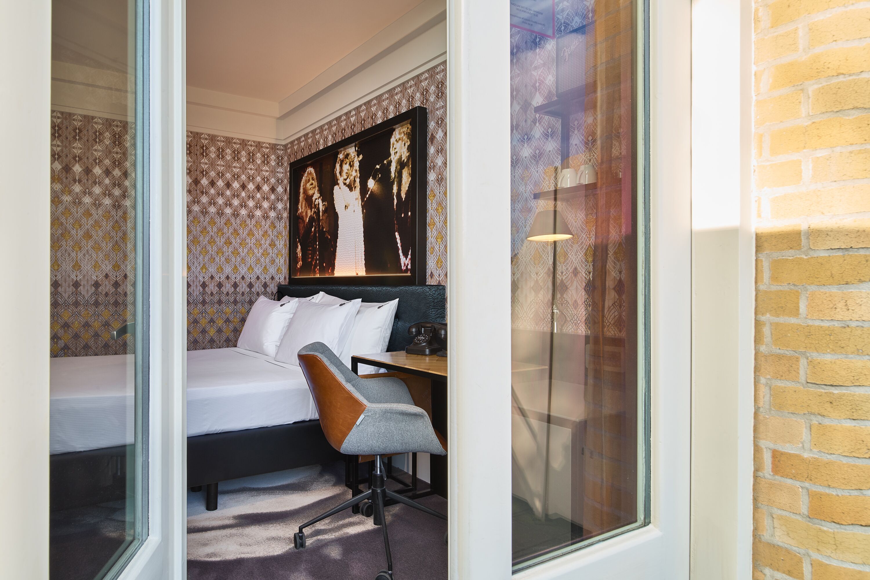 Een moderne hotelkamer met een bed, een bureau en een stoel. Er is een groot raam en een bakstenen muur.