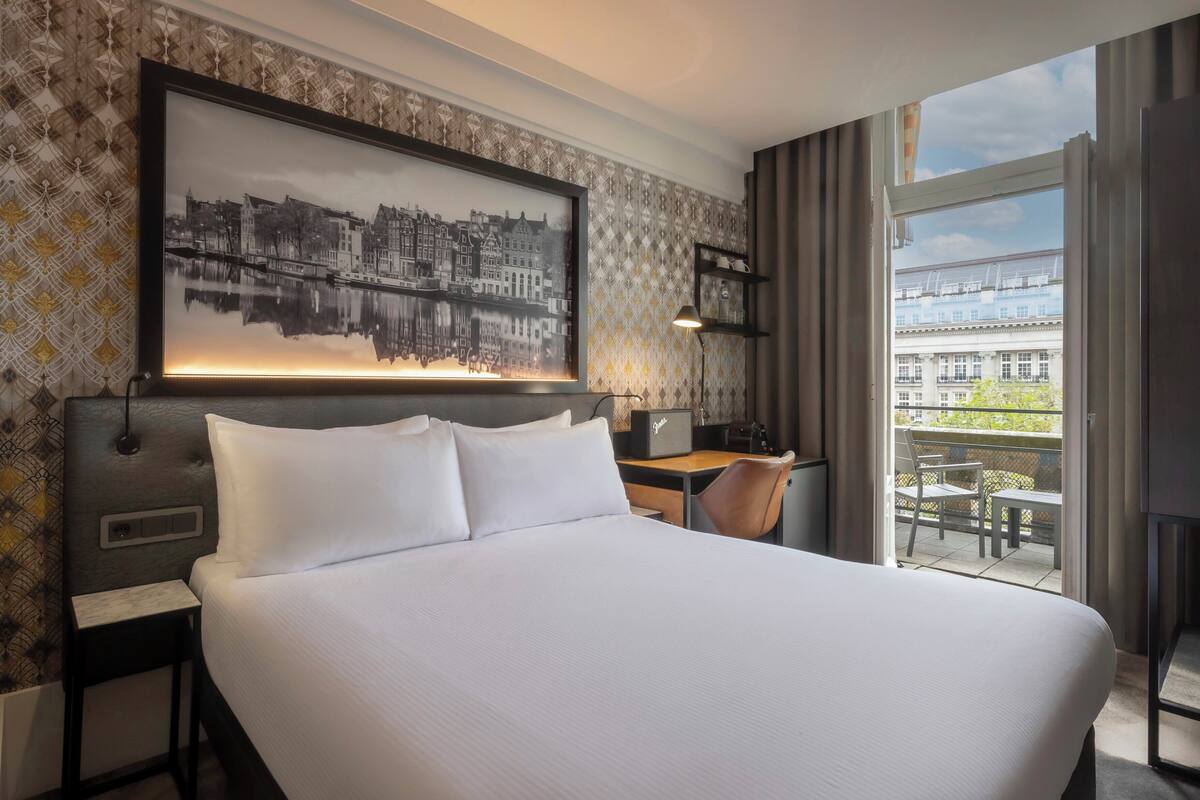 Une chambre d’hôtel avec un grand lit, un bureau avec une chaise, une petite table, une lampe et une grande fenêtre donnant sur des immeubles.