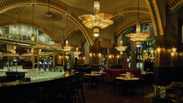 Un área de bar con varias mesas y sillas, una barra con vasos y botellas, y arcos de techo ornamentados con lámparas colgantes.