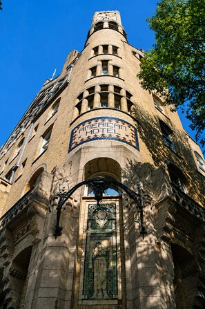 A historic building with a prominent clock tower, a mosaic sign reading 'AMERICAN HOTEL,' and a detailed stained glass window.