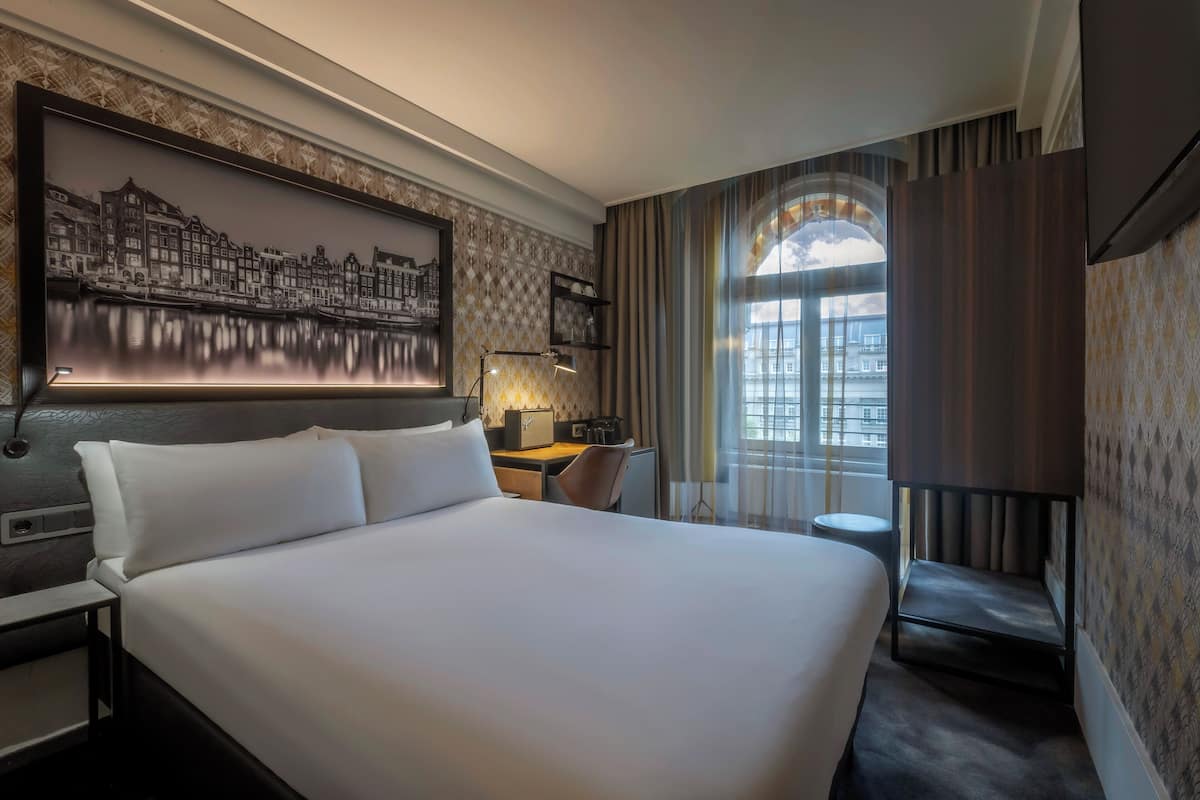 Une chambre d’hôtel avec un grand lit, un bureau avec une lampe, une chaise, une petite table et une grande fenêtre avec des rideaux.