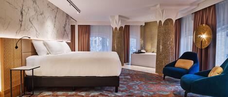 Una habitación de hotel moderna con una cama grande, una zona de estar con dos sillones, una mesita y una alfombra decorativa.