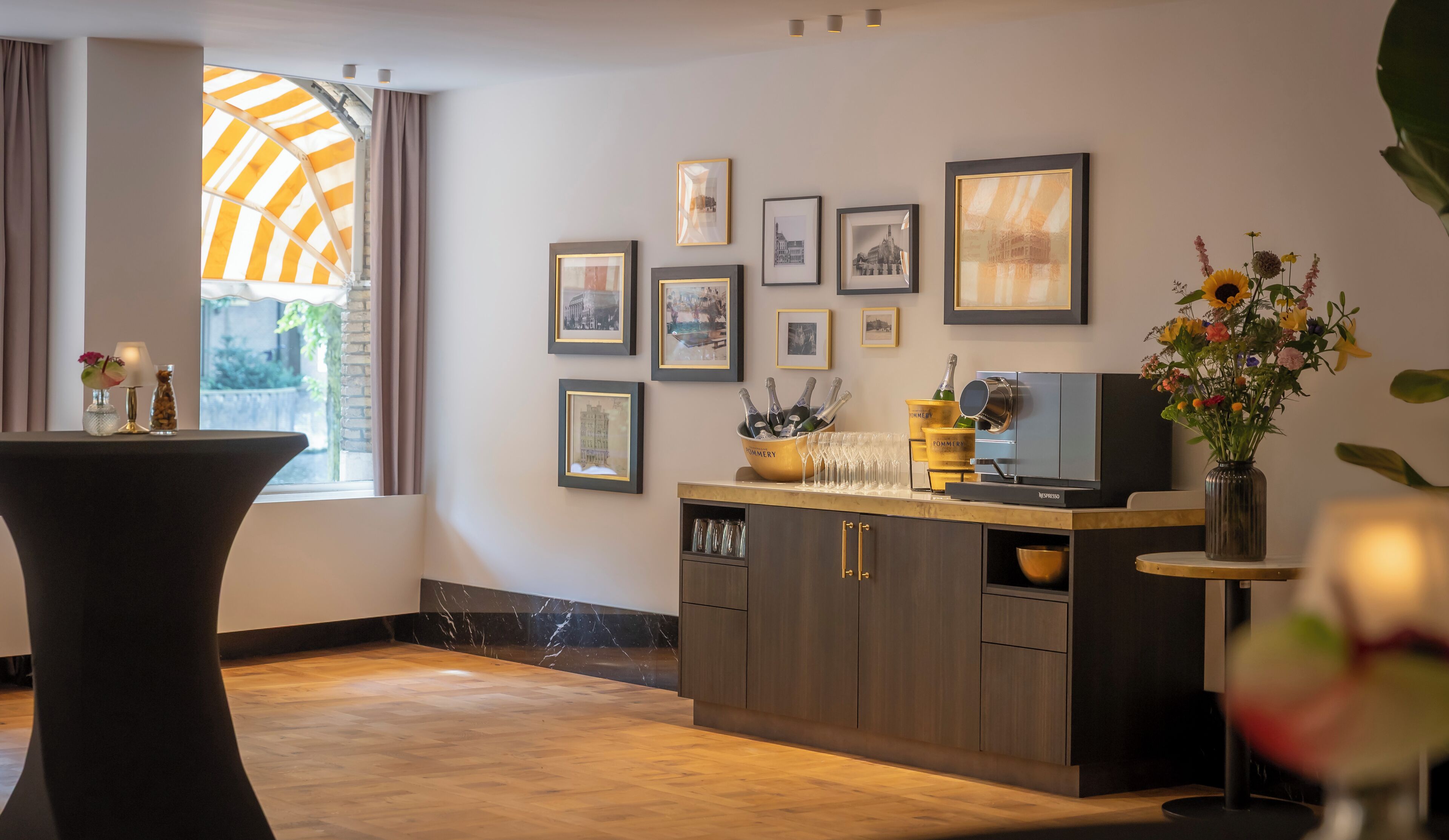 A modern interior with a wooden floor, a bar area with a Nespresso machine, framed pictures on the wall, and a window with curtains.