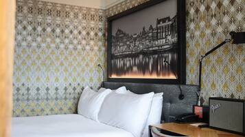 Habitación de hotel con cama, escritorio con lámpara, cuadro enmarco de un paisaje urbano y papel tapiz estampado.