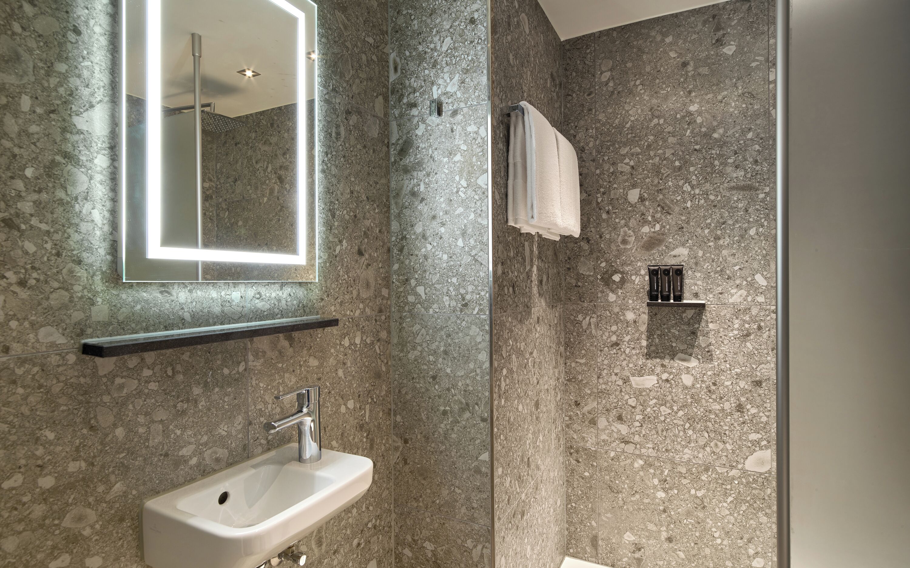 Un baño moderno con pared de ducha de granito, un lavabo blanco y un espejo con marco iluminado.