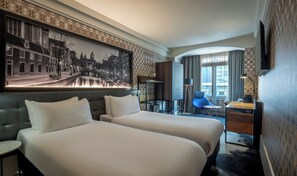 A hotel room with two beds, a desk, a chair, a large cityscape painting, and a view of the city through the window.