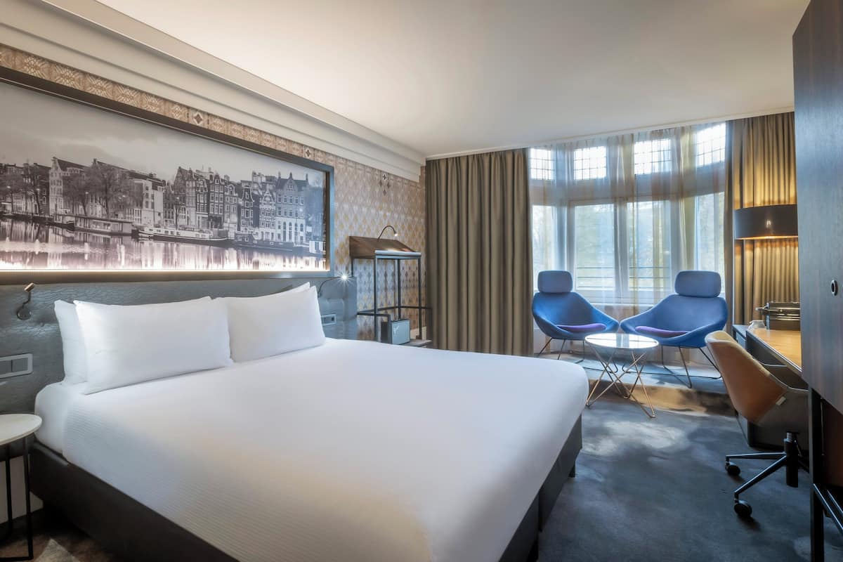 Une chambre d’hôtel avec un grand lit, deux chaises bleues, une petite table, un bureau, une lampe et un tableau représentant un paysage urbain accroché au mur.