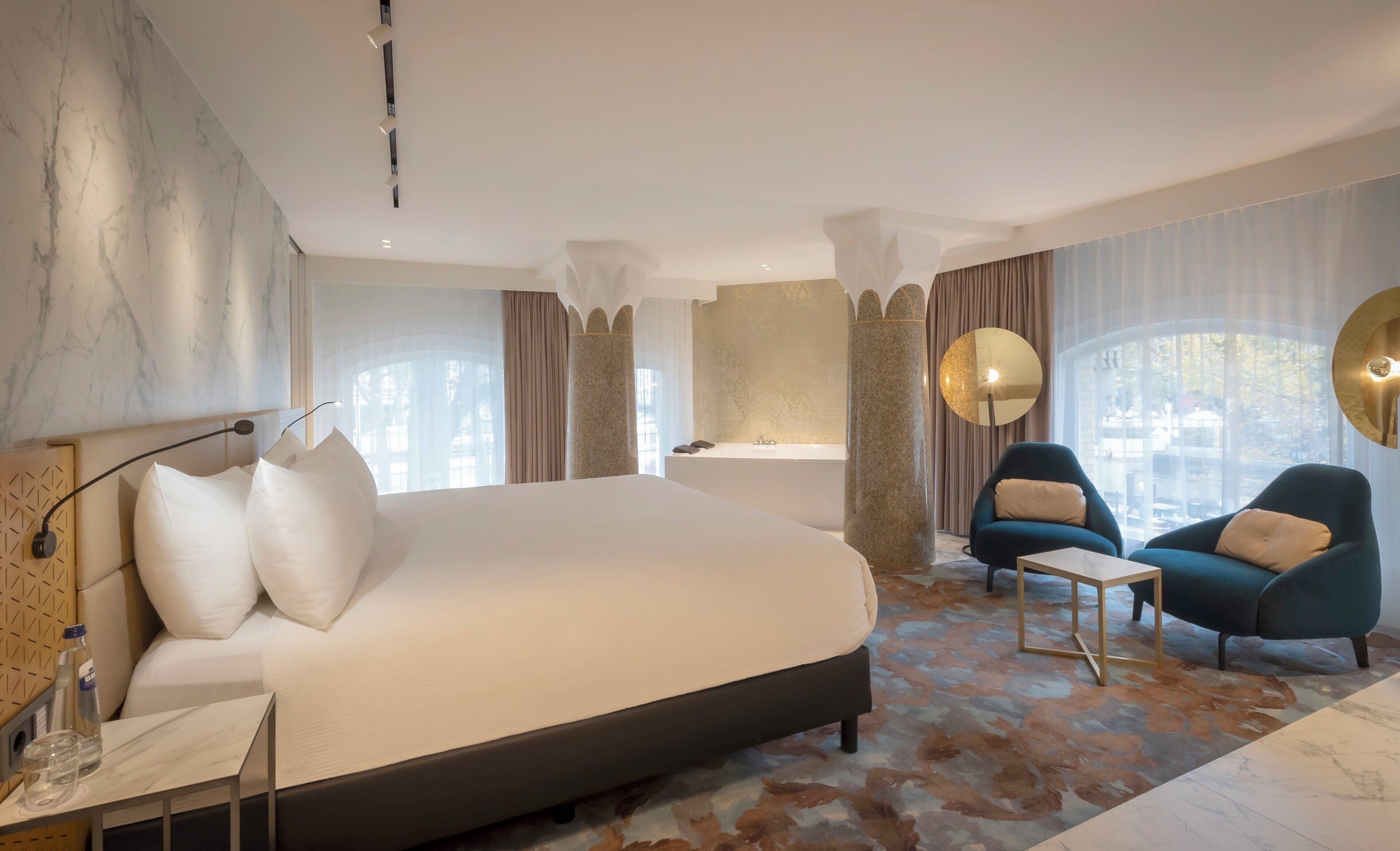 A modern hotel room with a large bed, a sitting area with two chairs, a bathtub, and a large window offering a view of the outdoors.