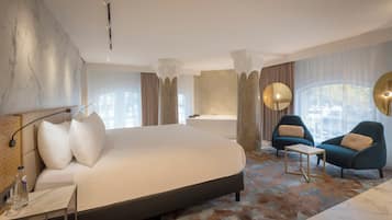 Habitación de hotel moderna con una cama grande, una zona de estar con dos sillones, una bañera y un ventanal con vistas al exterior.