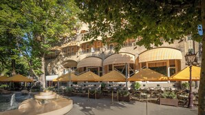 Ein Außenbereich eines Cafés mit mehreren gelb-weiß gestreiften Sonnenschirmen, einem Springbrunnen und Terrassenmöbeln.