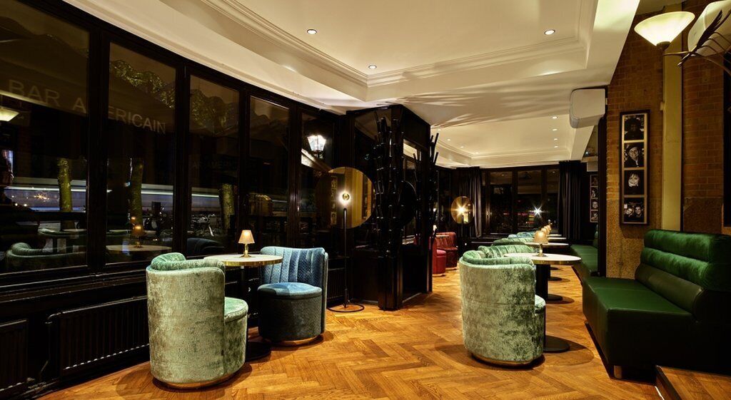 A bar area with round tables and cushioned seating, a piano, and a view through large windows.