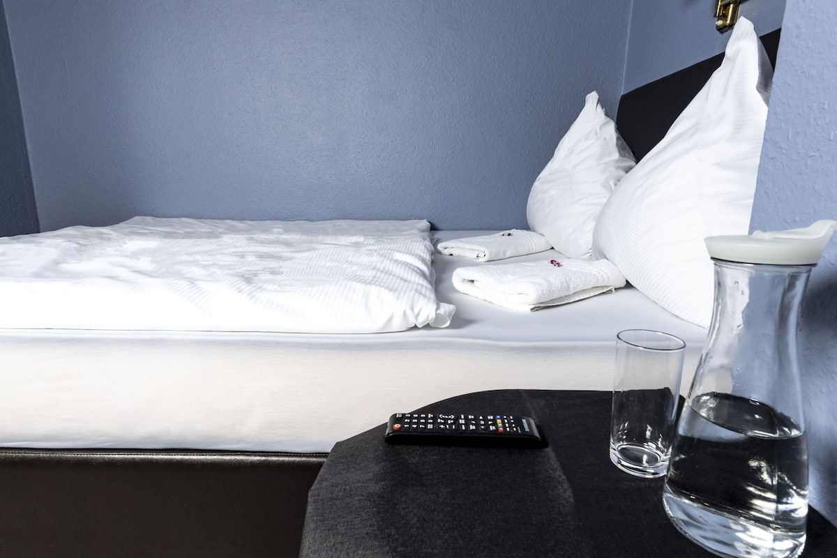 A neatly made bed with white linens, a remote control, a glass of water, and a pitcher on a bedside table in a room with blue walls.