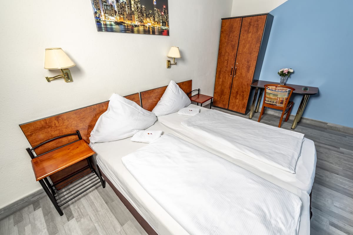 A neatly made bed with white linens, a wooden nightstand, a small table with a vase, and a cityscape painting on the wall.