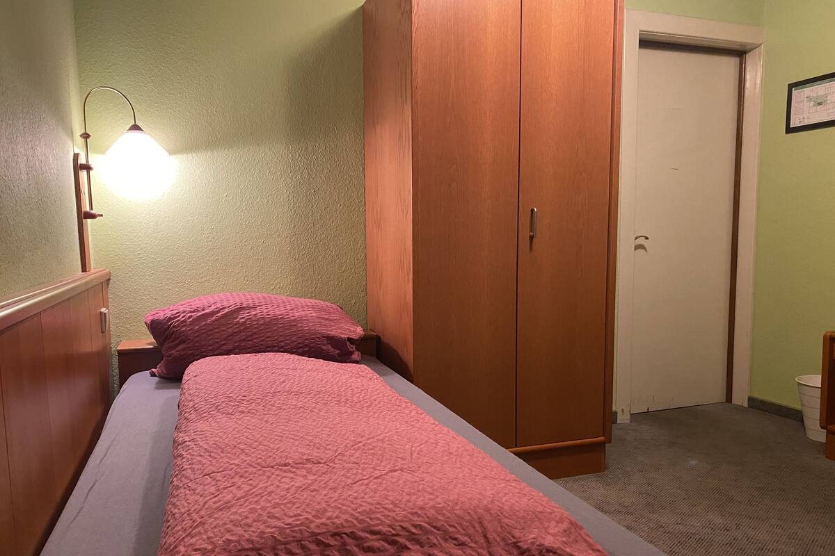 A single bed with a pink bedspread, a wooden wardrobe, and a nightstand with a lamp.
