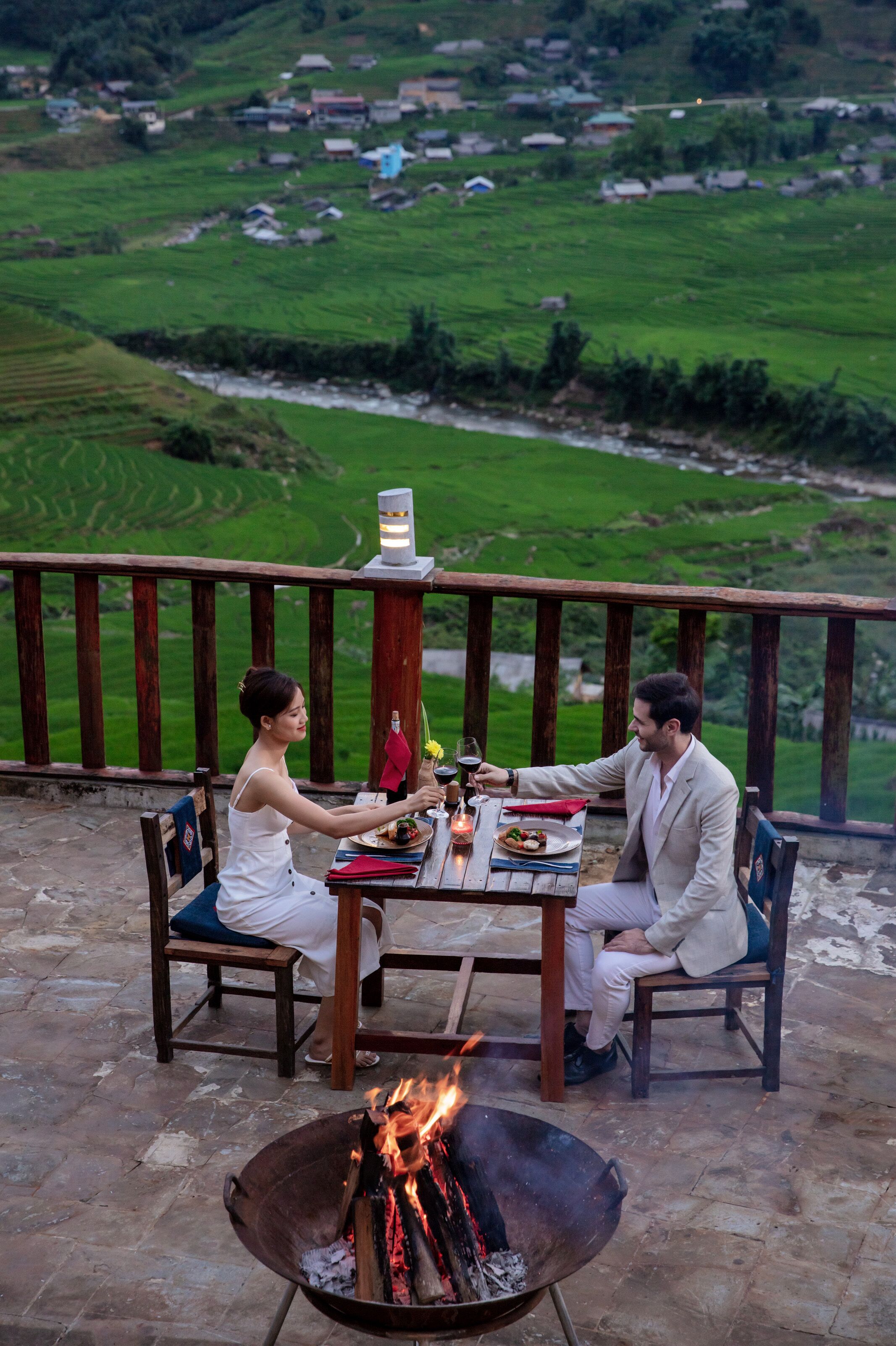 A couple dining outdoors with a fire pit in the foreground and a scenic view of a green valley in the background.