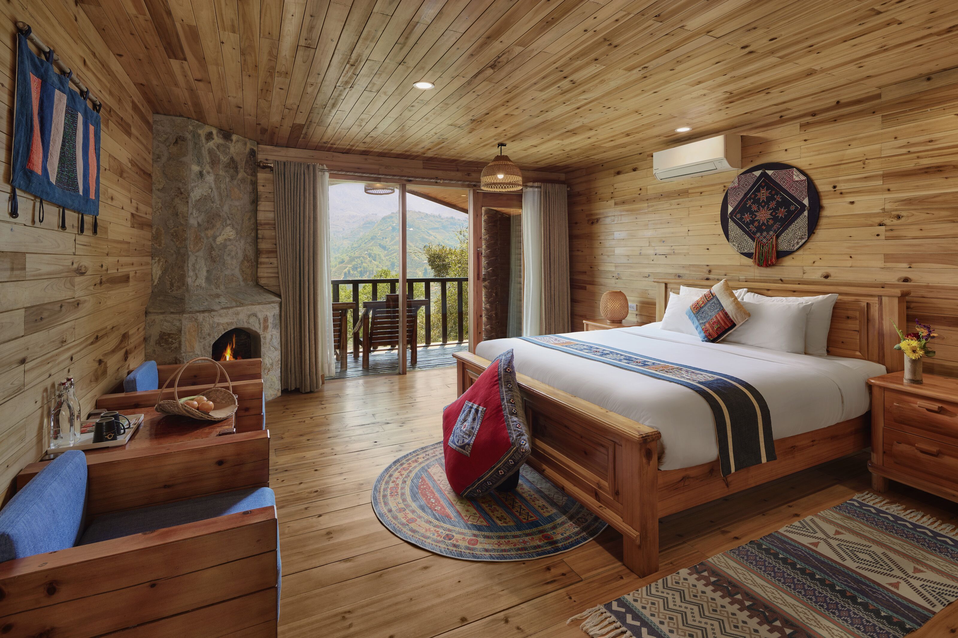 A wooden cabin bedroom with a stone fireplace, a bed with a patterned blanket, a wooden nightstand, and a view of the outdoors.