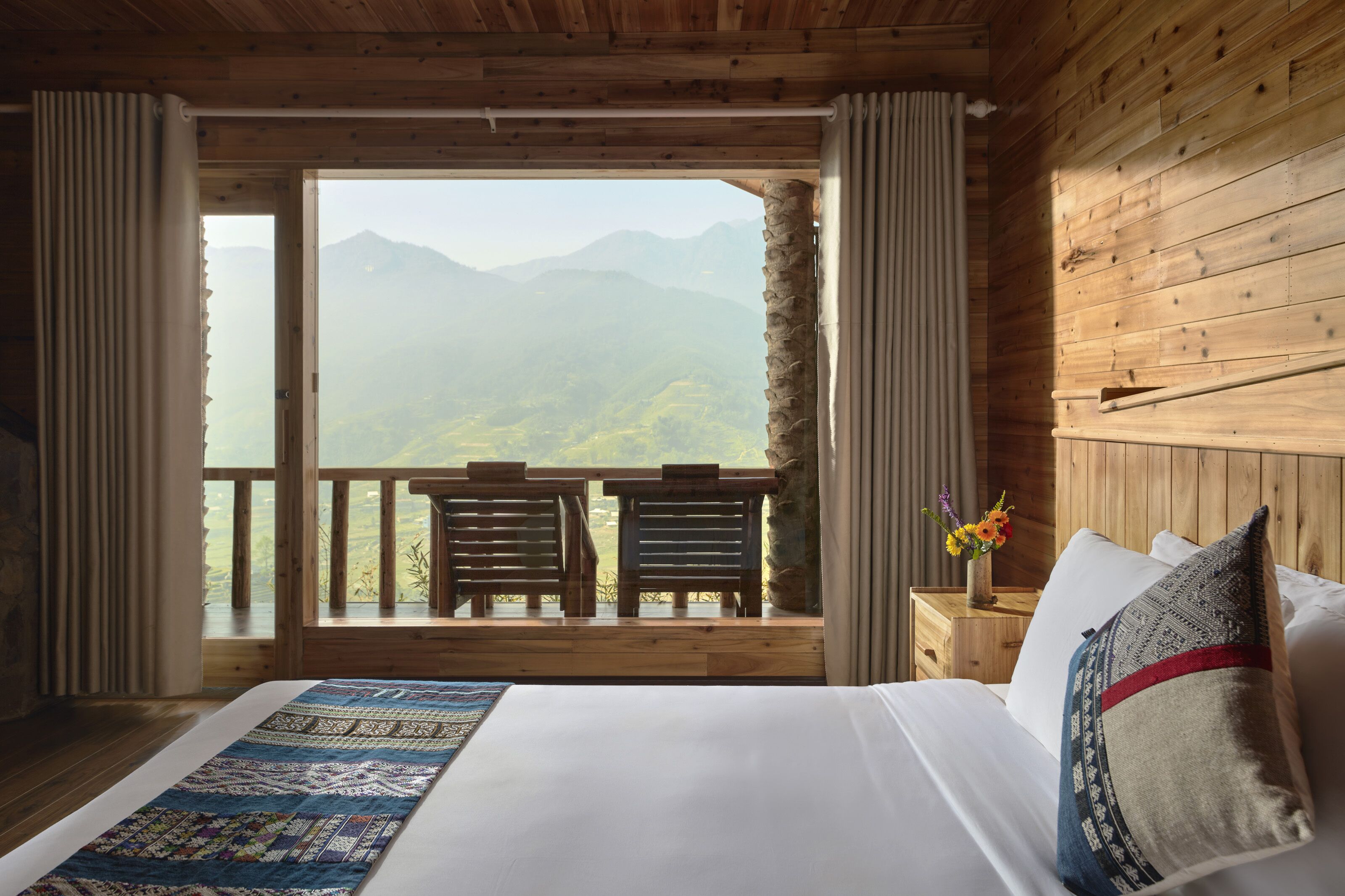A bedroom with a large bed, wooden walls, a balcony with two chairs, and a view of mountains.